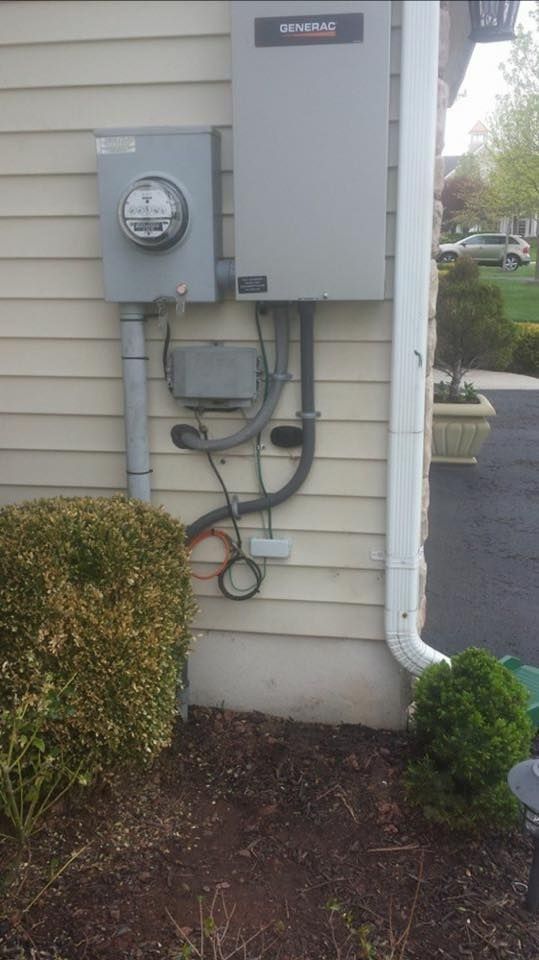 Electrical meter box and equipment mounted on a beige siding exterior wall near landscaping and a downspout.