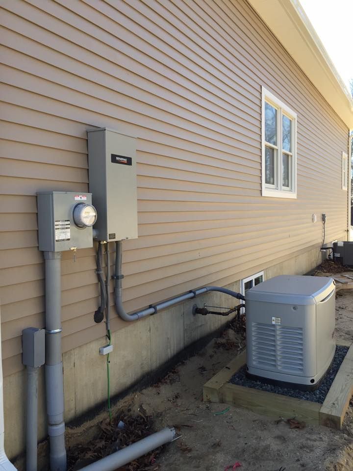 Electrical equipment on a beige house: meter box, generator, and conduits. Gray boxes and pipes against tan siding.