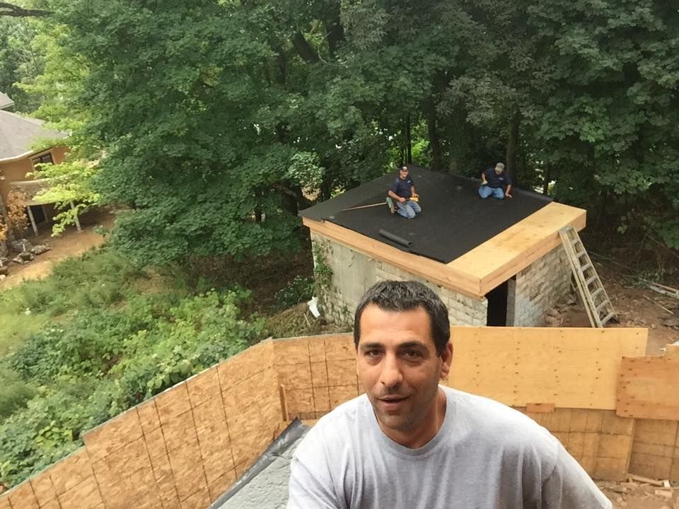 Man smiling, construction workers on a small roof. Building under construction, green trees in background.