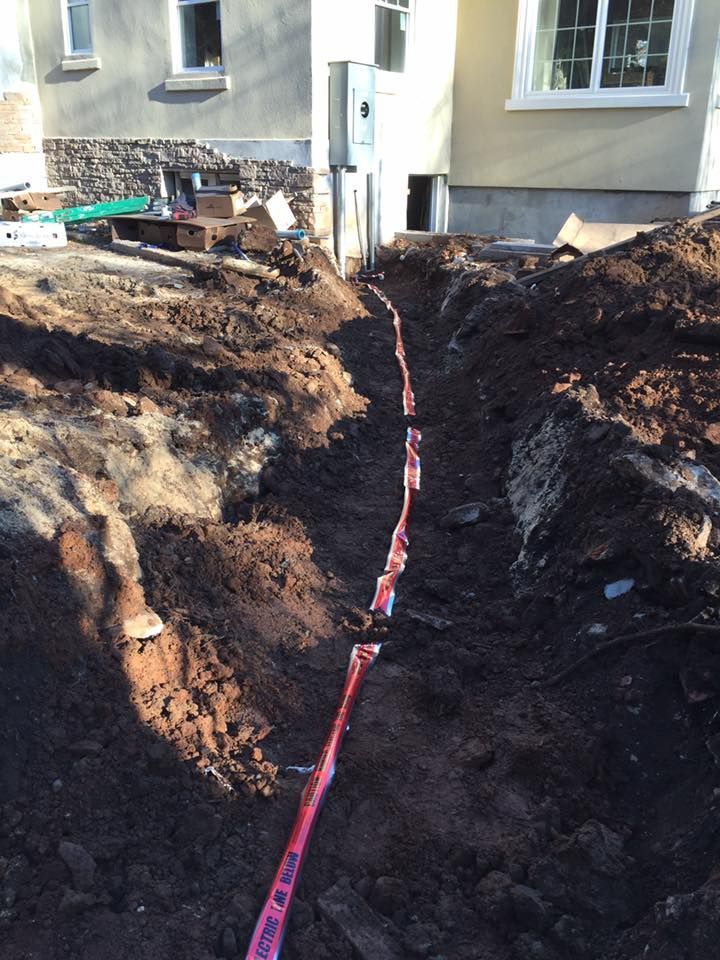 Trench dug in dark soil with red tape marking a utility line, alongside a building.