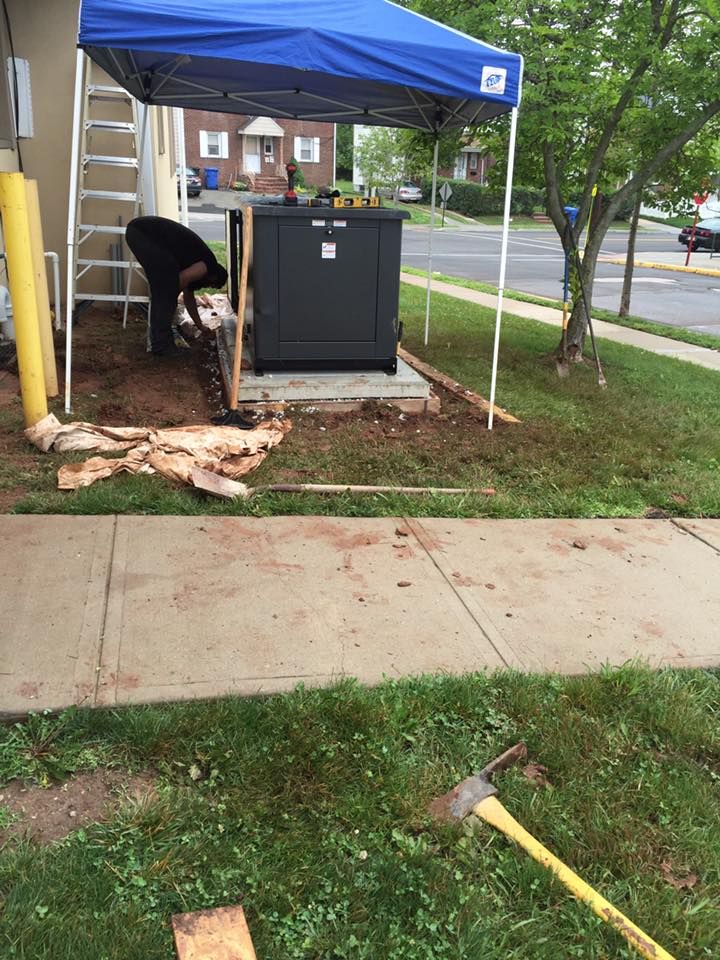 Person working on a black electrical box outside, under a blue canopy. Located on a concrete pad near a sidewalk and grass.