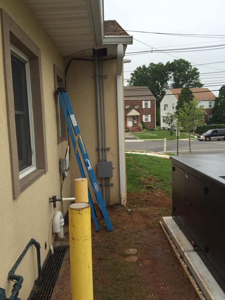 A blue ladder leaning against a building with electrical conduits. Yellow poles and a generator are also present.