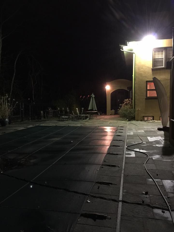 Nighttime backyard scene with a pool covered in a tarp, a building with lights, and trees in the background.