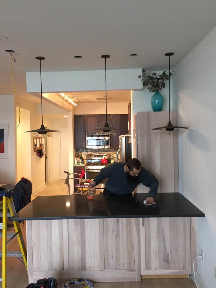 Man cleaning a dark countertop in a kitchen with pendant lights.