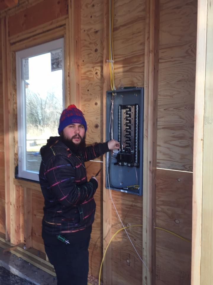 Man pointing at electrical panel in a wood-framed building, wearing a beanie and jacket, indoors.