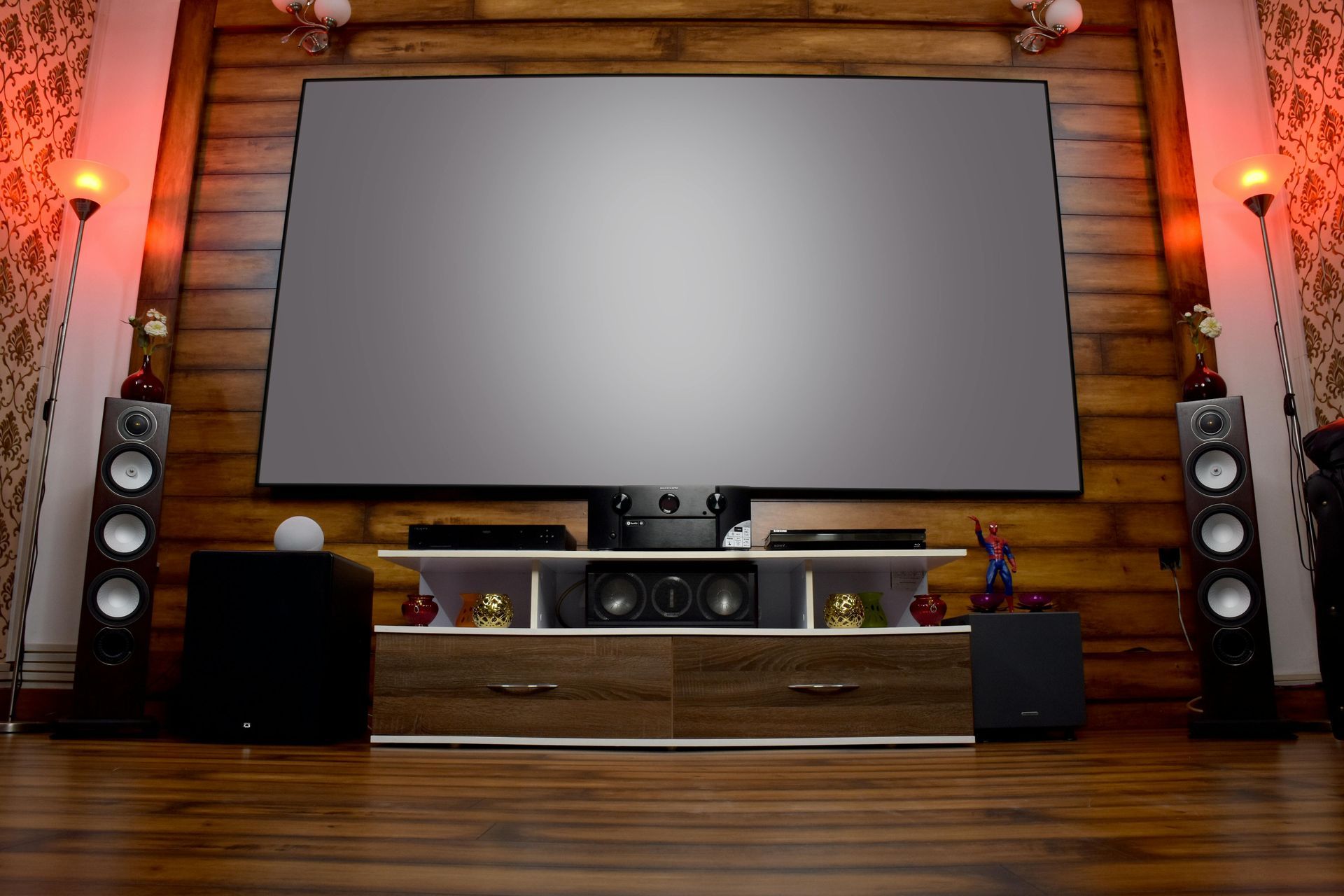 Home theater setup with large screen, speakers, and media console against a wood-paneled wall, warm lighting.