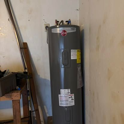 A gray water heater is sitting in a corner of a room next to a wooden table.