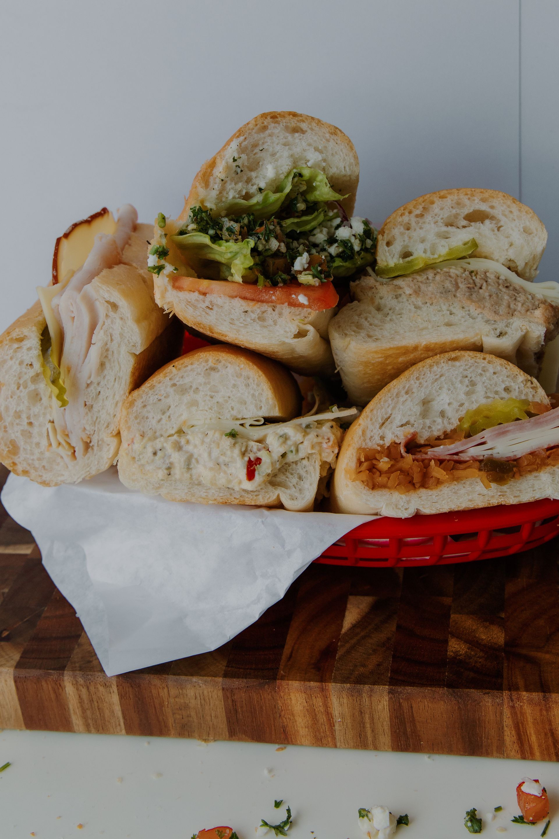 Basket of assorted sandwiches, piled on white paper, on a wooden surface.