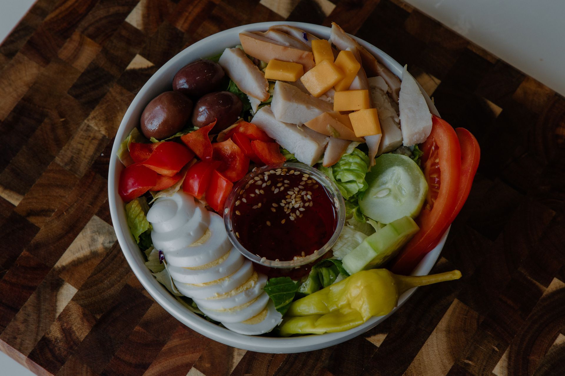 Salad bowl with chicken, cheese, olives, red pepper, cucumber, pepperoncini, egg, and dressing on a wooden board.