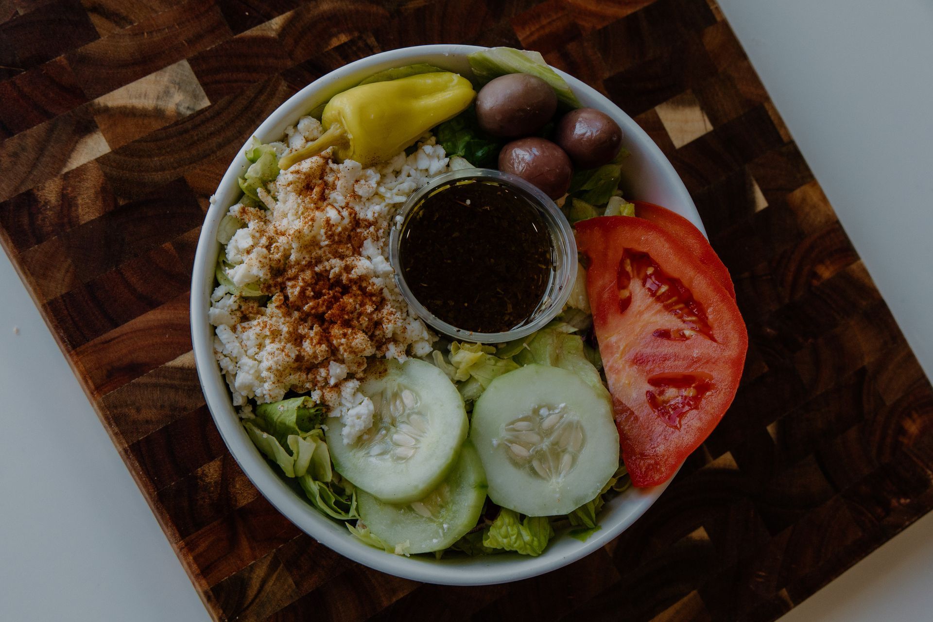 Greek salad in a white bowl, with cucumbers, tomatoes, olives, and dressing on a wooden board.