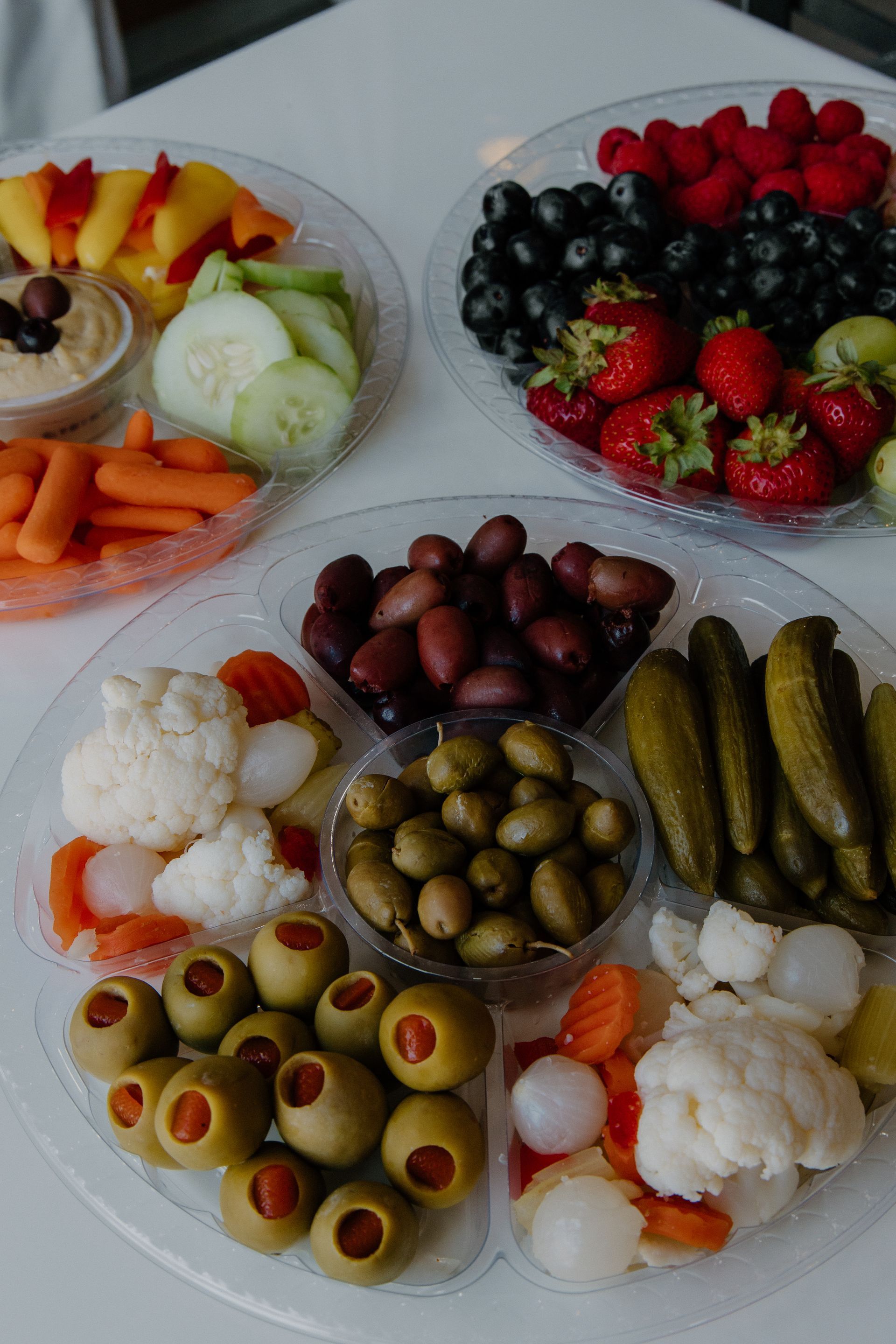 Arrangement of various fruits, vegetables, and olives in clear platters on a white surface.