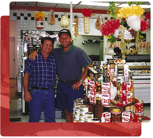 Two men in a deli, one with arm around the other. Surrounded by food products, hanging decorations, and deli counter.