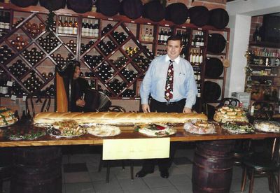 Man in restaurant with large sandwich, surrounded by food, wine bottles, and an accordion player.