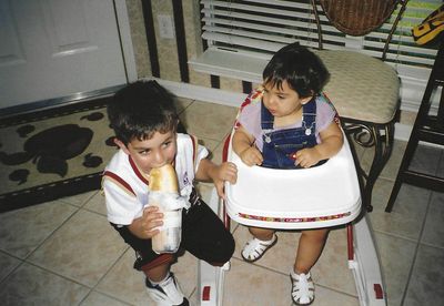 Two young children, one eating from bottle, other in walker. Indoors with tile floor, door, window.