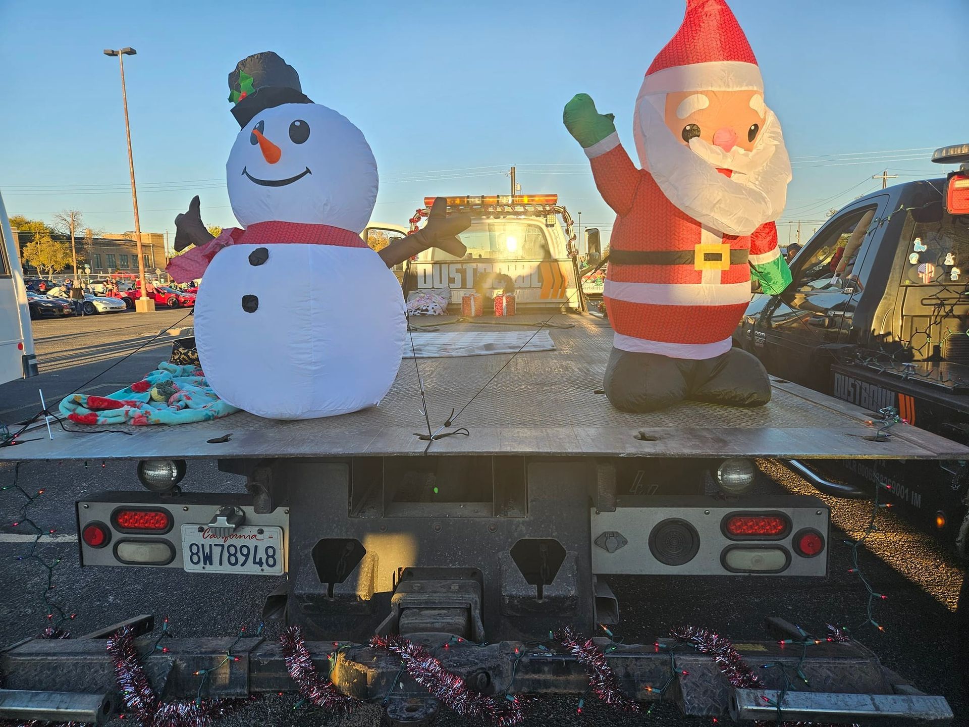 Inflatable snowman and Santa on a tow truck bed decorated with garland, set outdoors.