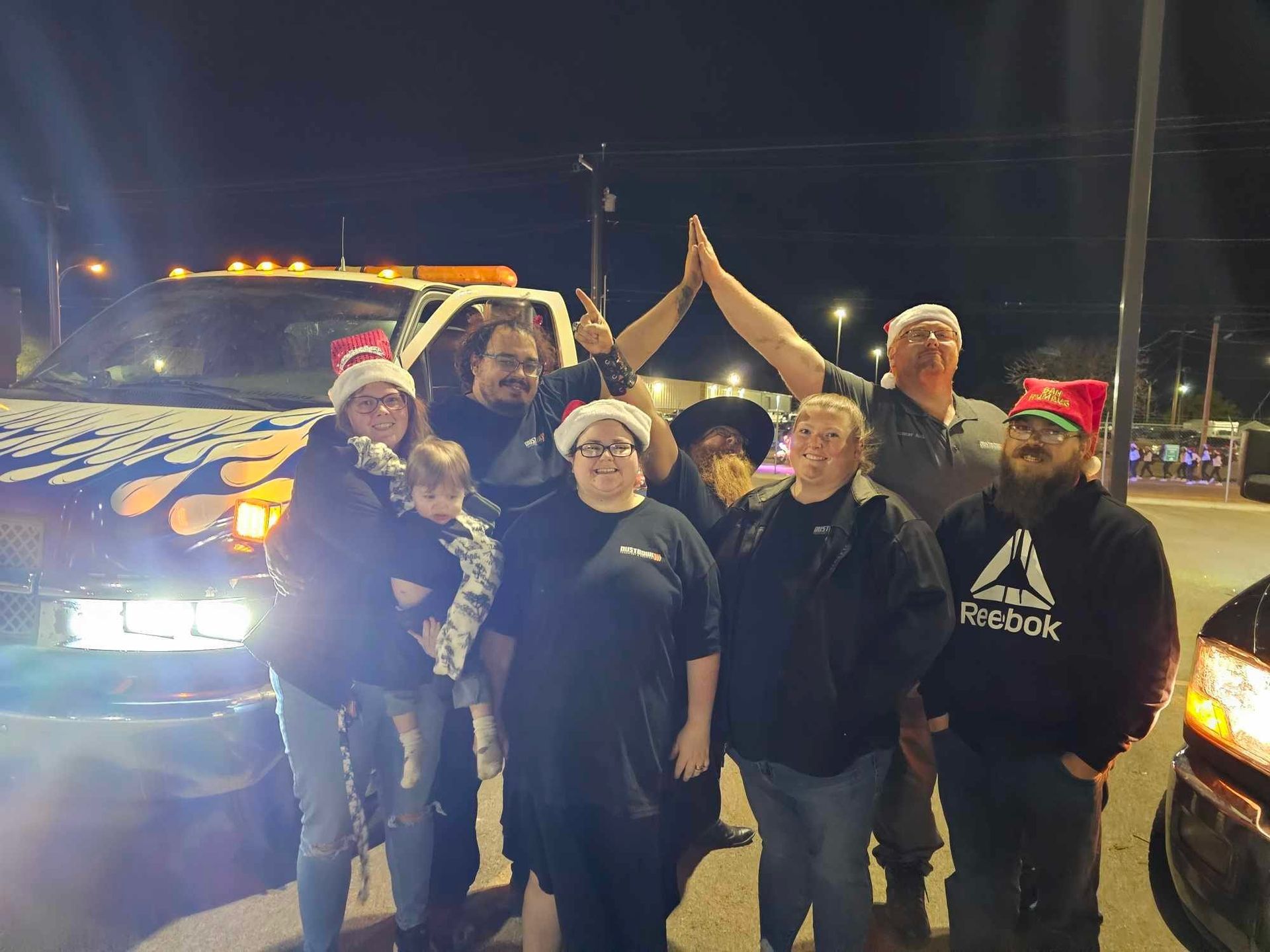 Group of people wearing Santa hats, pose in front of a truck with Christmas lights at night, smiling.