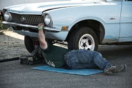 A person under a light blue car, working on it. On a mat, outside.