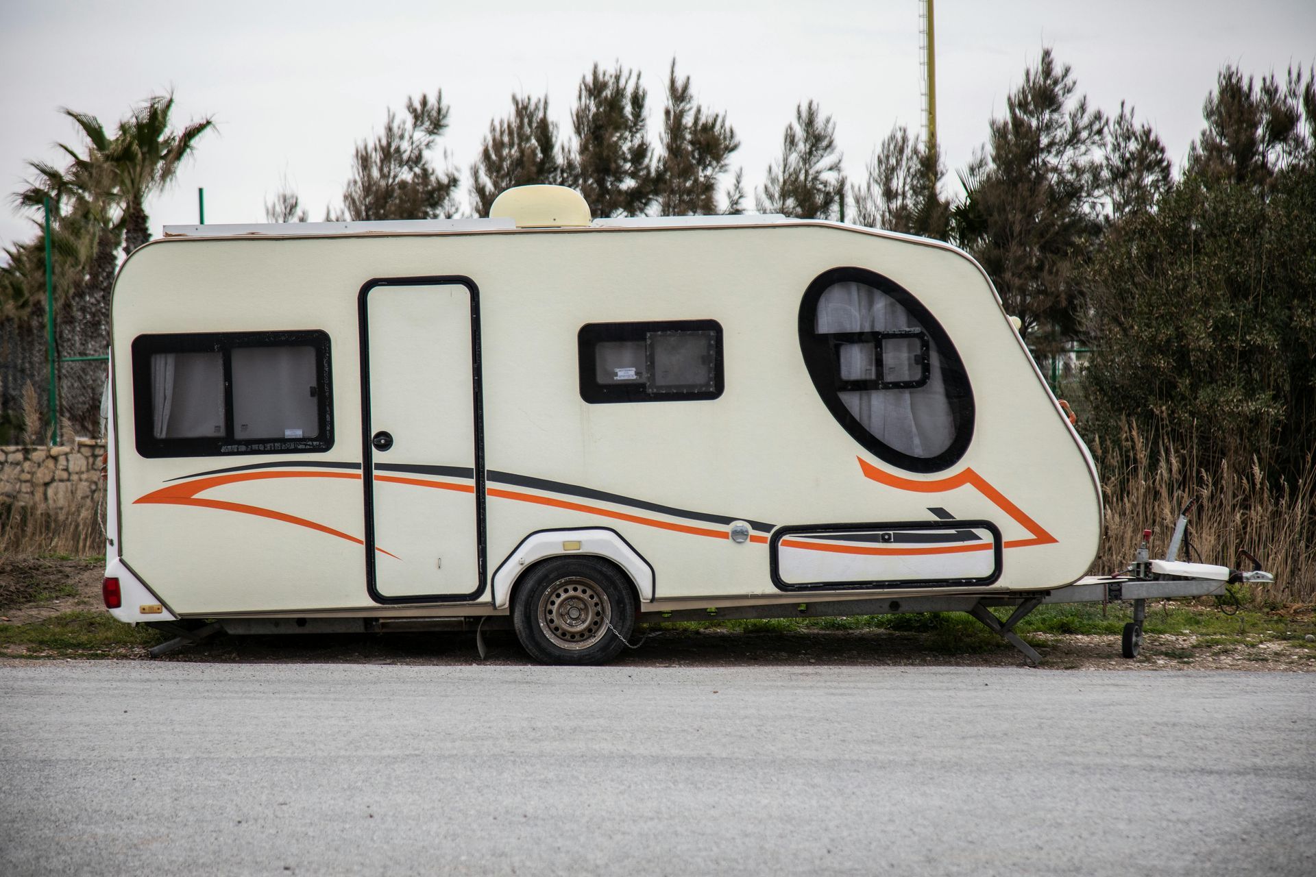 Cream-colored travel trailer parked on a paved surface with a black and orange design, trees in background.