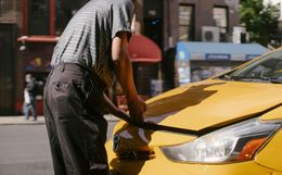 Person in a grey shirt and dark pants opens the hood of a yellow taxi on a city street.