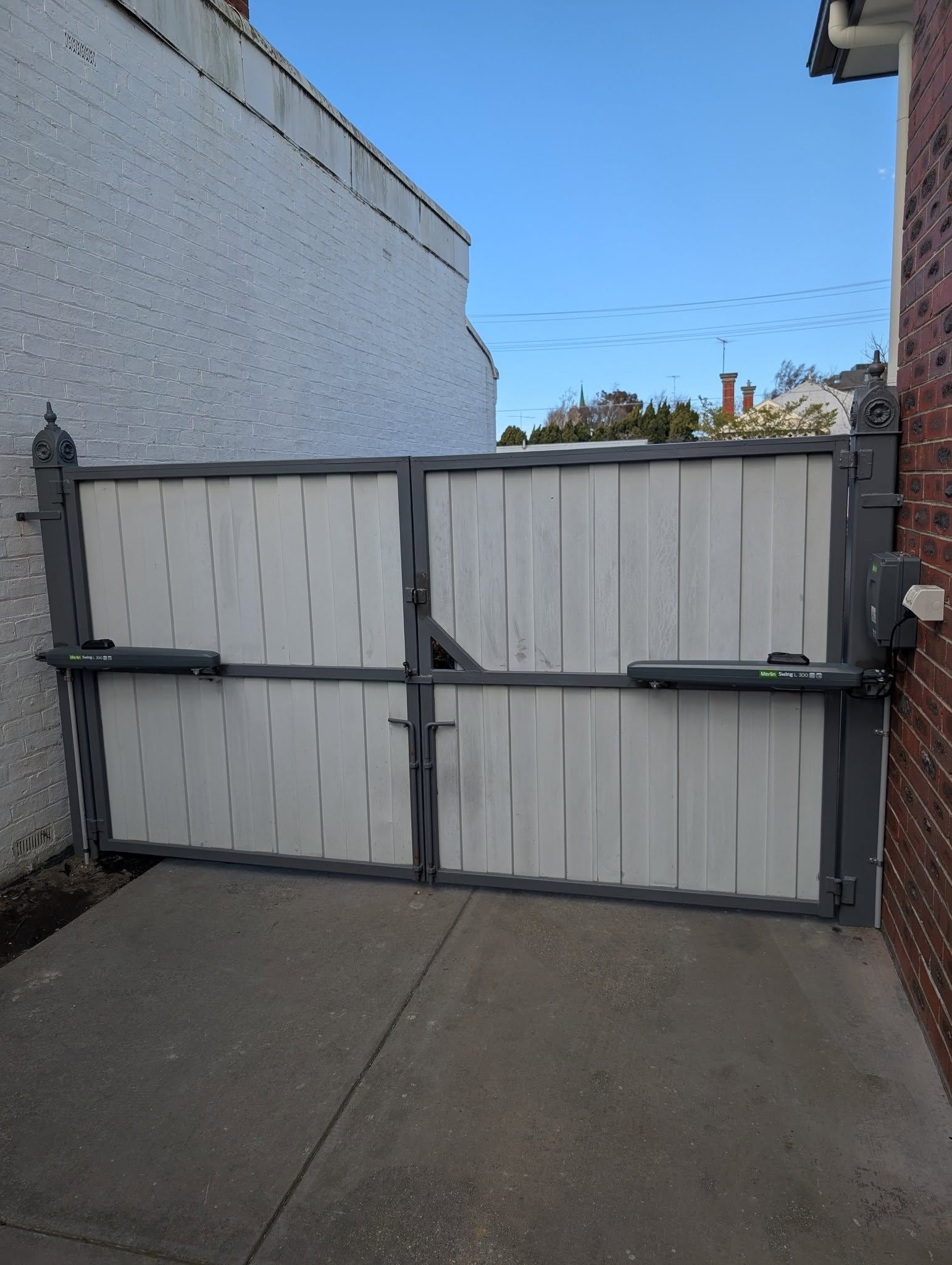 Gray Automatic Driveway Gate Between a White Building and a Brick Wall — Goldfields Garage Doors & Gates in Wendouree, VIC