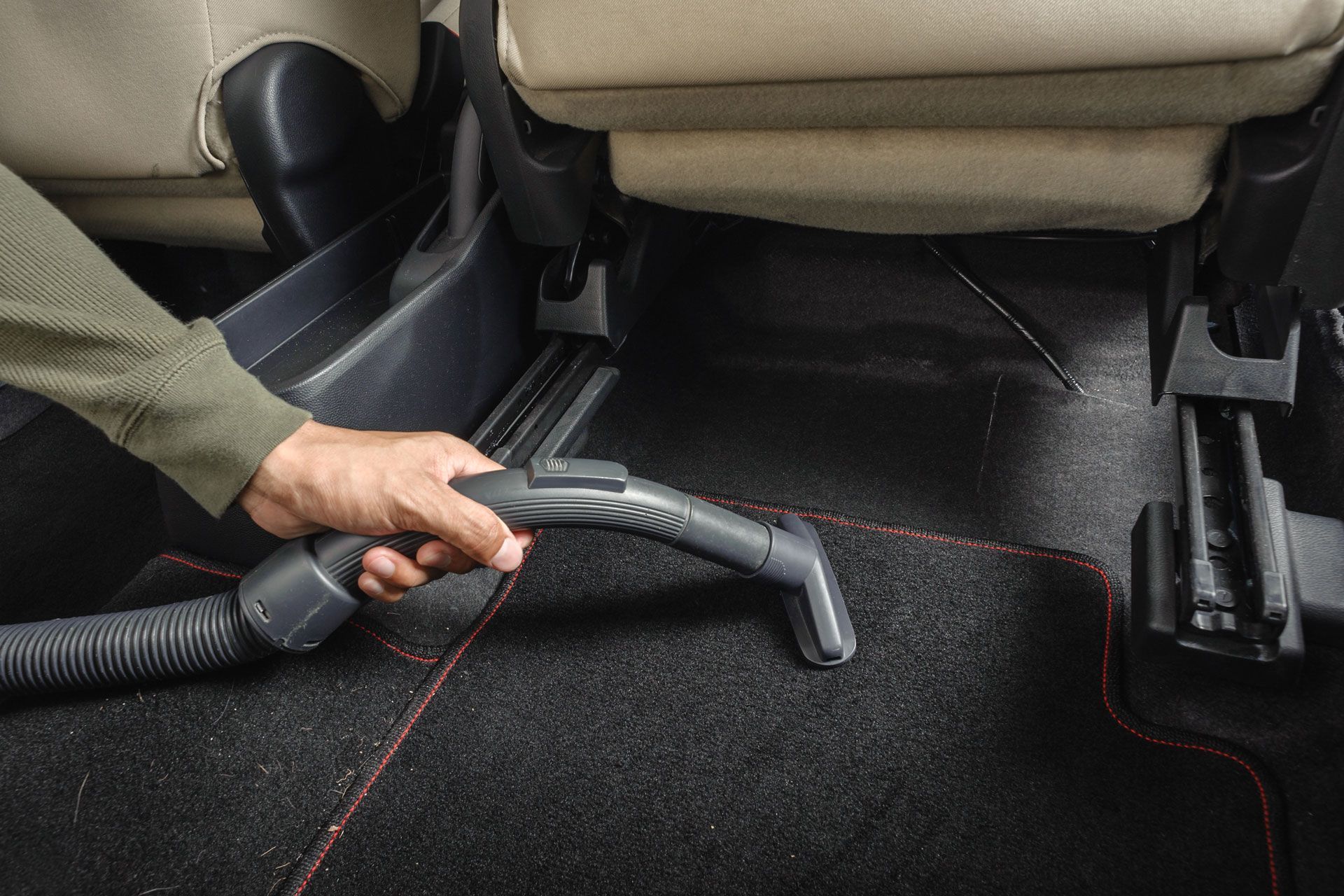 Person vacuuming the black carpeted floor of a car using a handheld vacuum attachment.