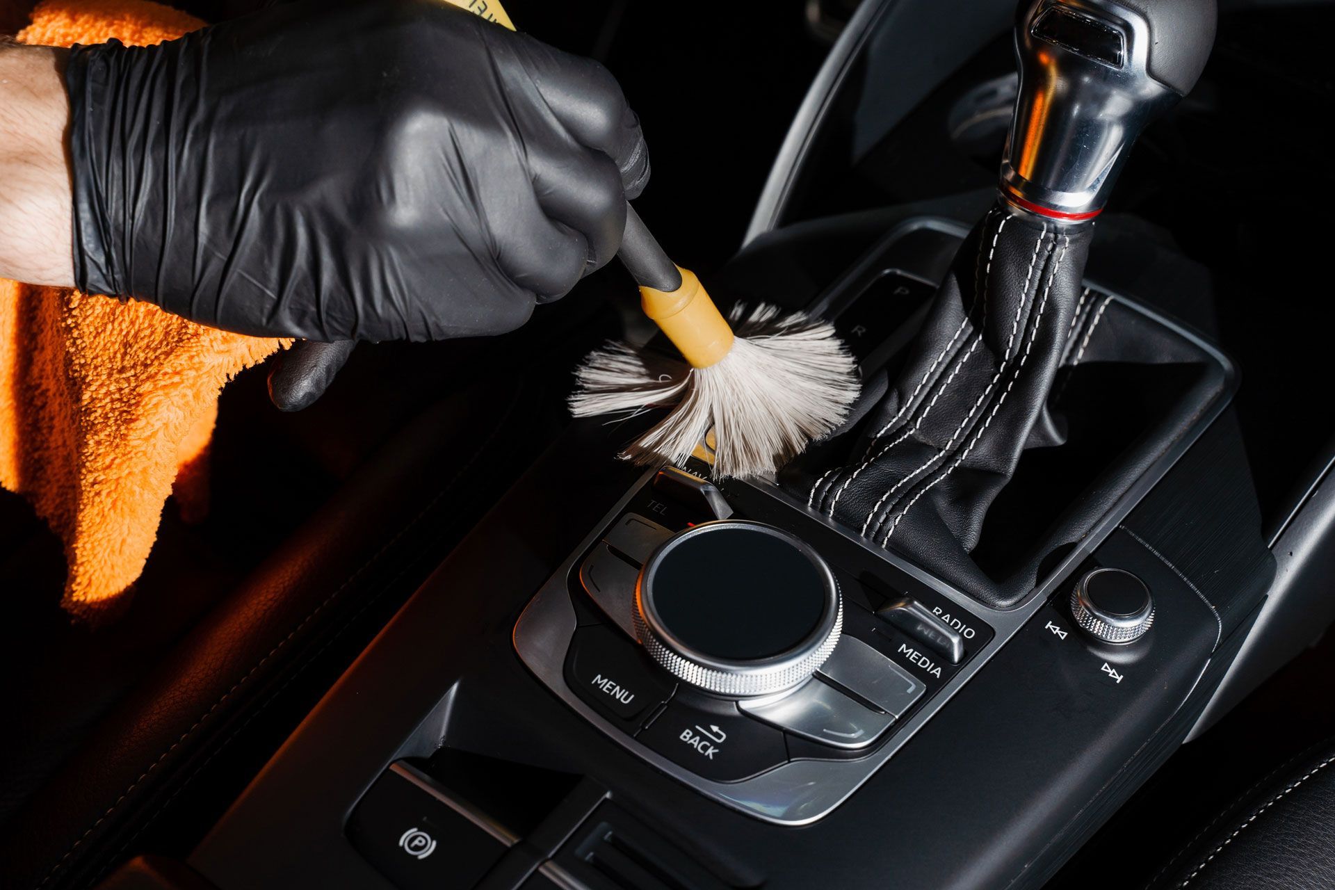 Black-gloved hand cleaning a car's center console with a brush; orange cloth in the background.