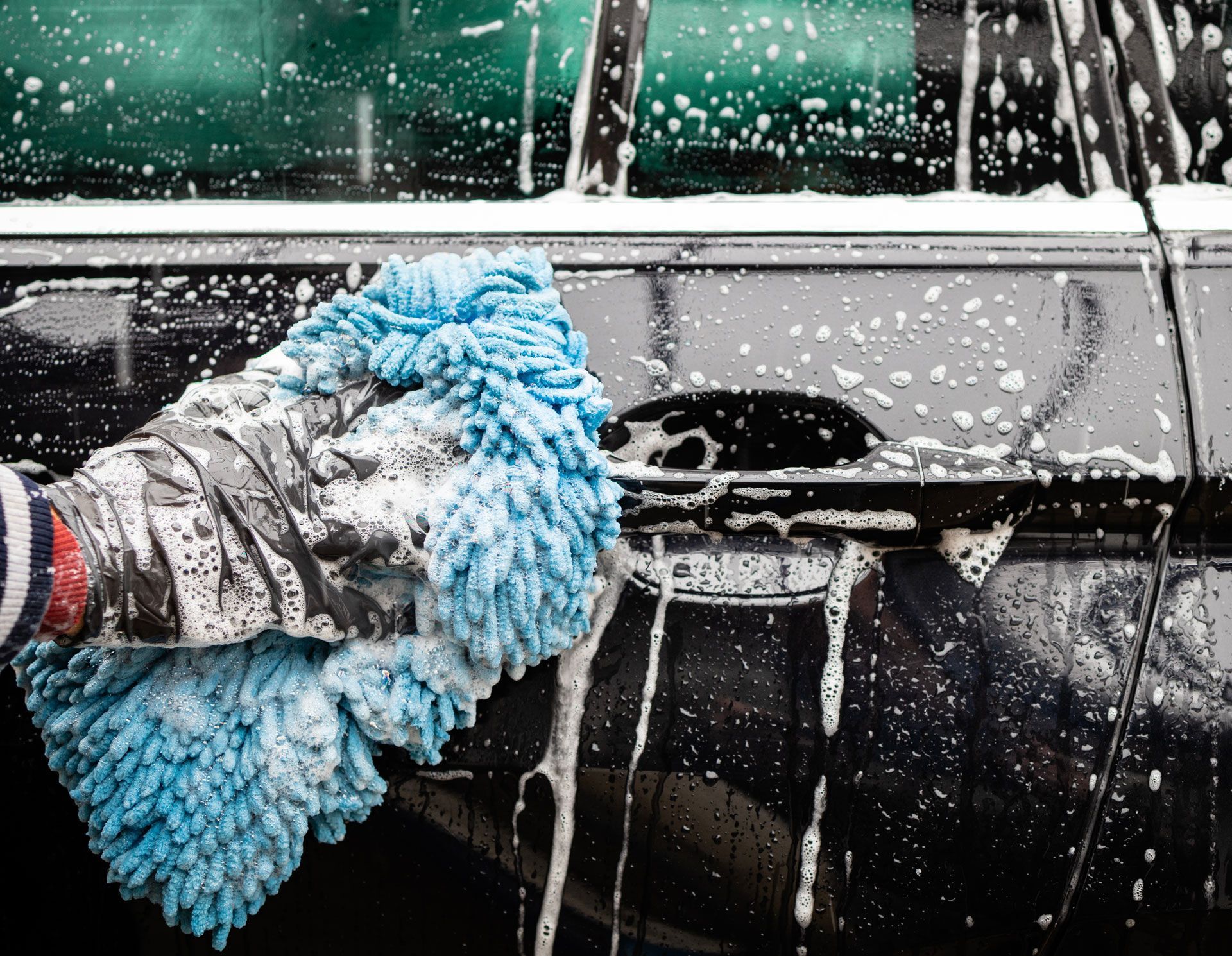 Person washing a black car with a blue microfiber mitt covered in soapy water.