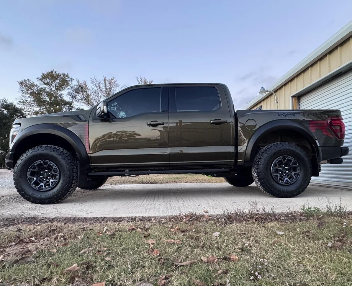 An olive-drab Ford F-150 Raptor pickup truck parked on a concrete driveway, featuring American flag decals.