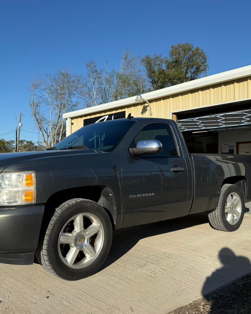 A gray single-cab Chevrolet Silverado pickup truck parked on a concrete lot in front of a metal warehouse.
