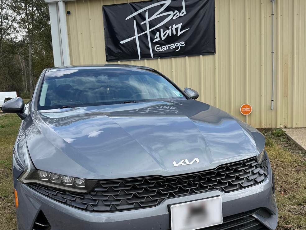 A gray Kia K5 sedan parked in front of a metal building with a 