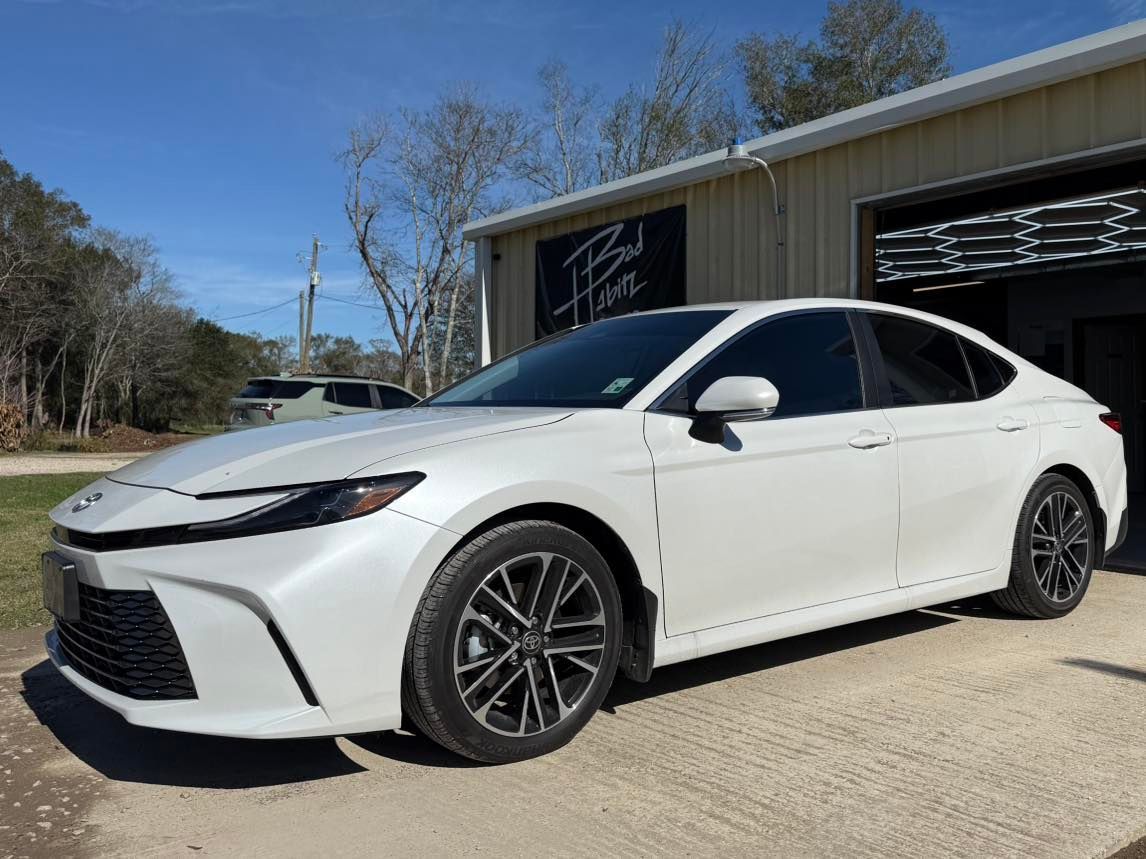 A white Toyota Camry sedan parked on a concrete lot in front of a building on a sunny day.
