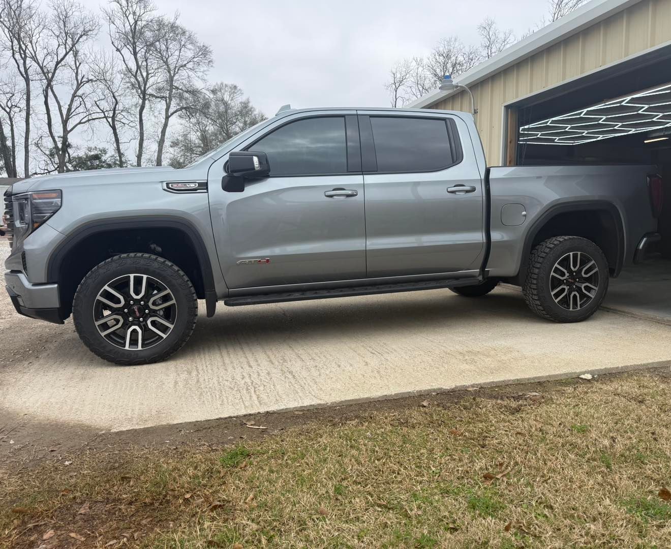 A silver GMC Sierra crew cab pickup truck parked on a concrete driveway in front of a metal building.
