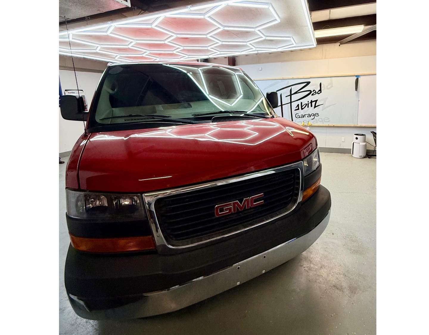 A bright red GMC Savana van parked inside a workshop with honeycomb-patterned overhead lighting.