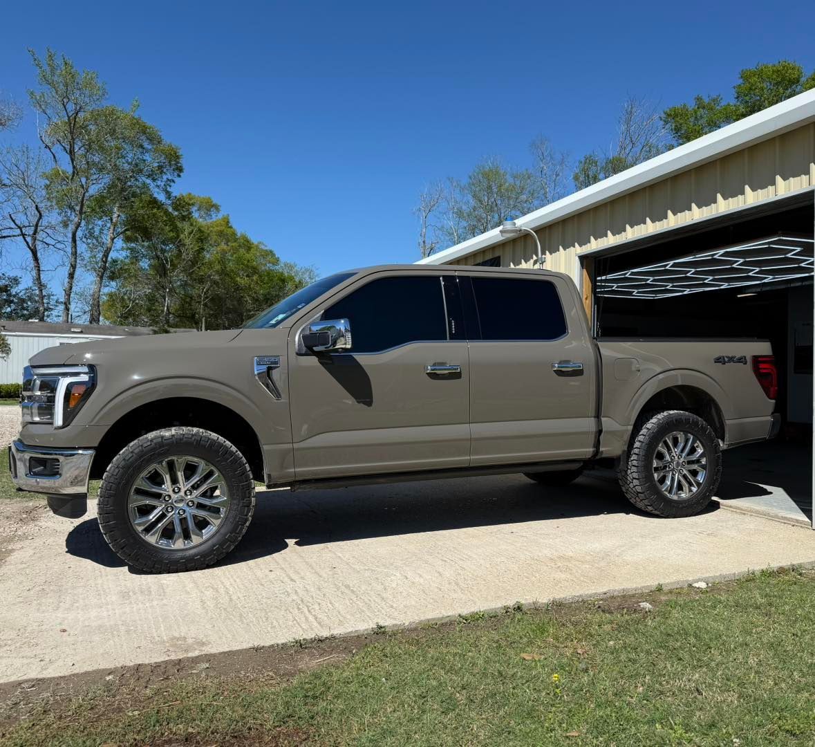 A tan crew cab Ford F-150 pickup truck parked on a concrete driveway next to a metal building under a clear blue sky.
