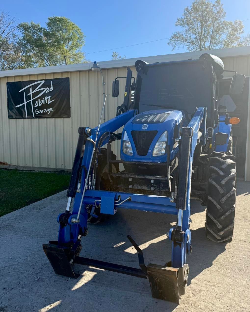 A blue New Holland tractor with a front-end loader parked on a concrete lot in front of a building with a black sign.