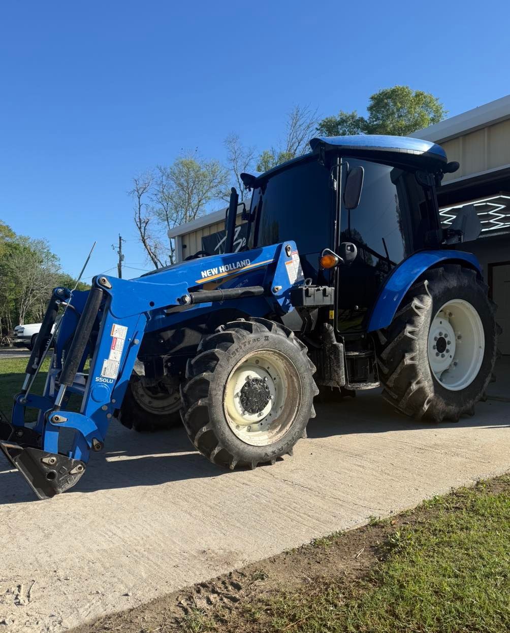 A blue tractor with a front-end loader parked on a concrete driveway outside a building on a sunny day.