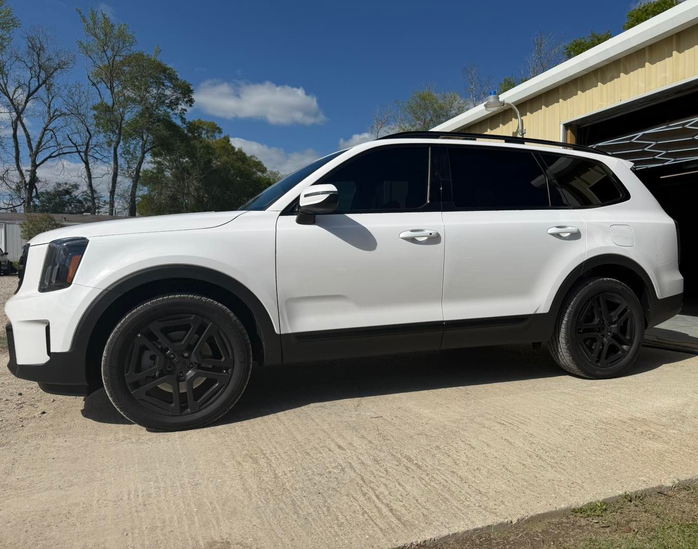 A white Kia Telluride SUV parked on a concrete driveway in front of a metal building on a sunny day.