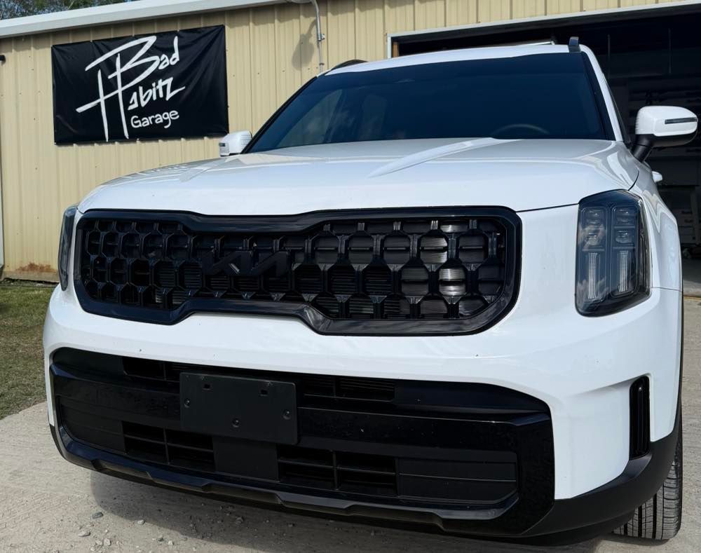 A white Kia Telluride with a custom black grille parked outside a building with a 
