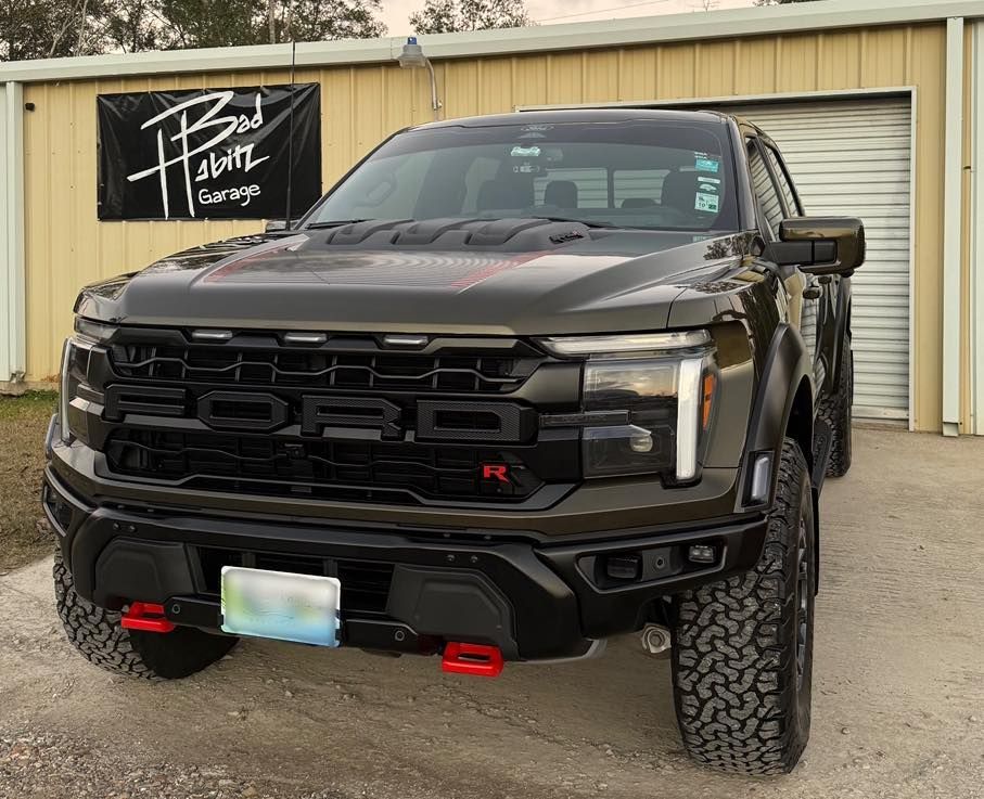 A dark bronze Ford F-150 Raptor R with red tow hooks parked in front of a tan metal building with a