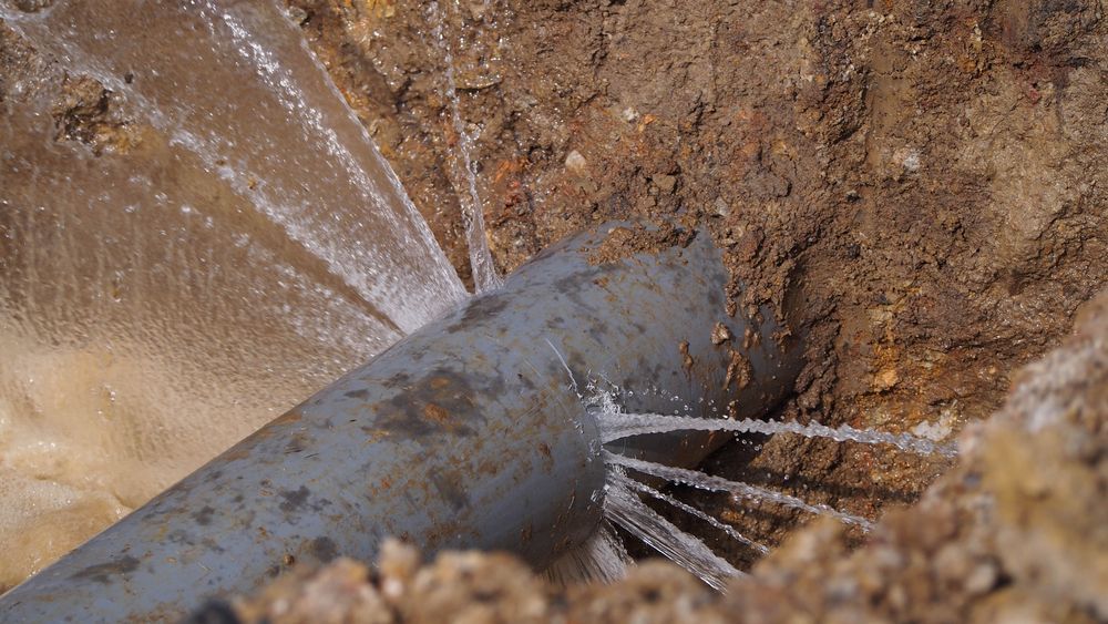 Leaking grey pipe in a brown dirt trench, with water spraying out. — Darwin Plumbing Maintenance in Knuckey Lagoon, NT