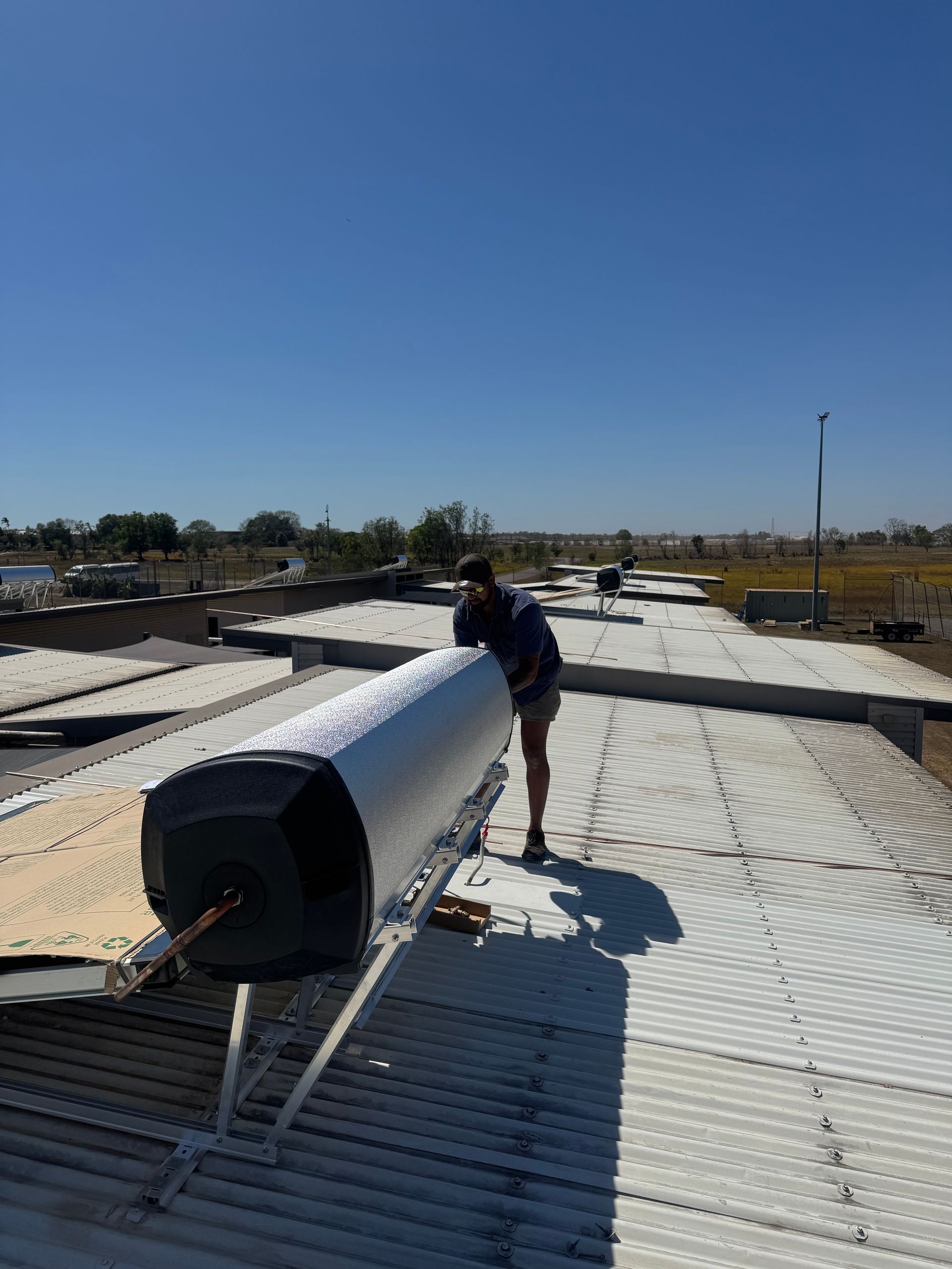 Person installing a solar water heater on a rooftop under a clear blue sky. — Darwin Plumbing Maintenance in Knuckey Lagoon, NT