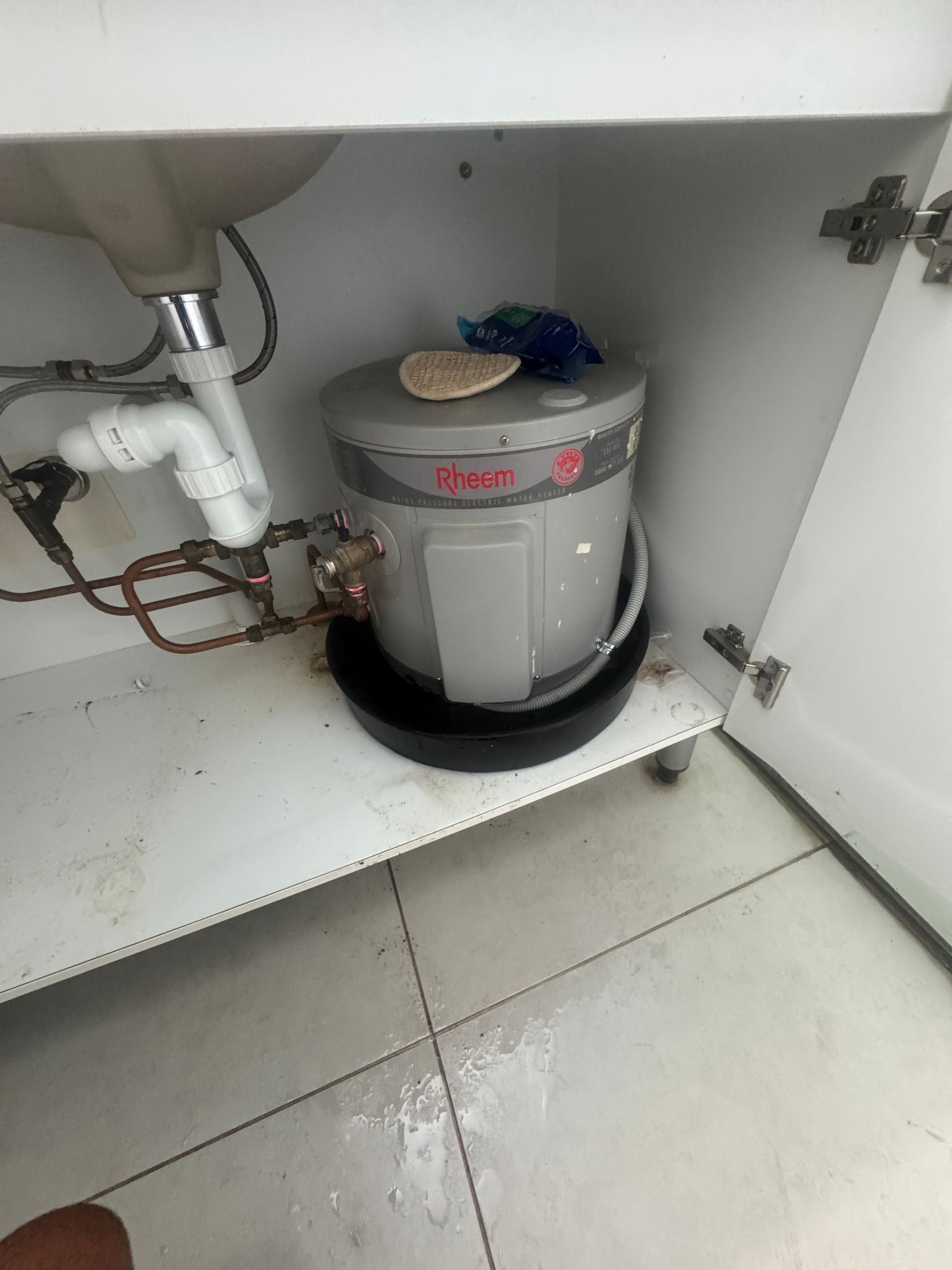 A small water heater under a sink in a white cabinet. Gray tank with black base, copper pipes, and white plumbing. — Darwin Plumbing Maintenance in Knuckey Lagoon, NT