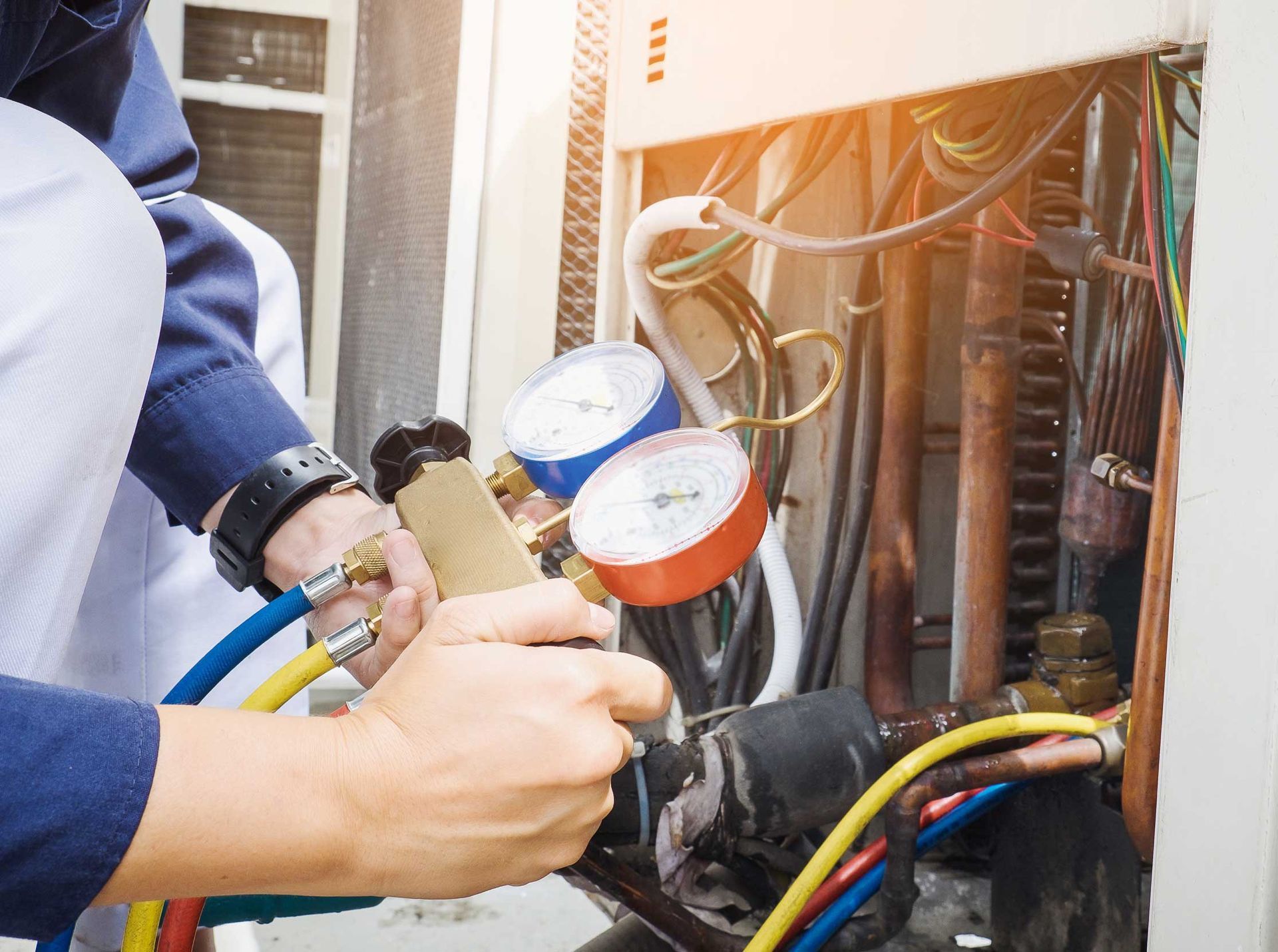 HVAC technician using gauges to service an air conditioning unit; hands, hoses, dials, and wires visible.