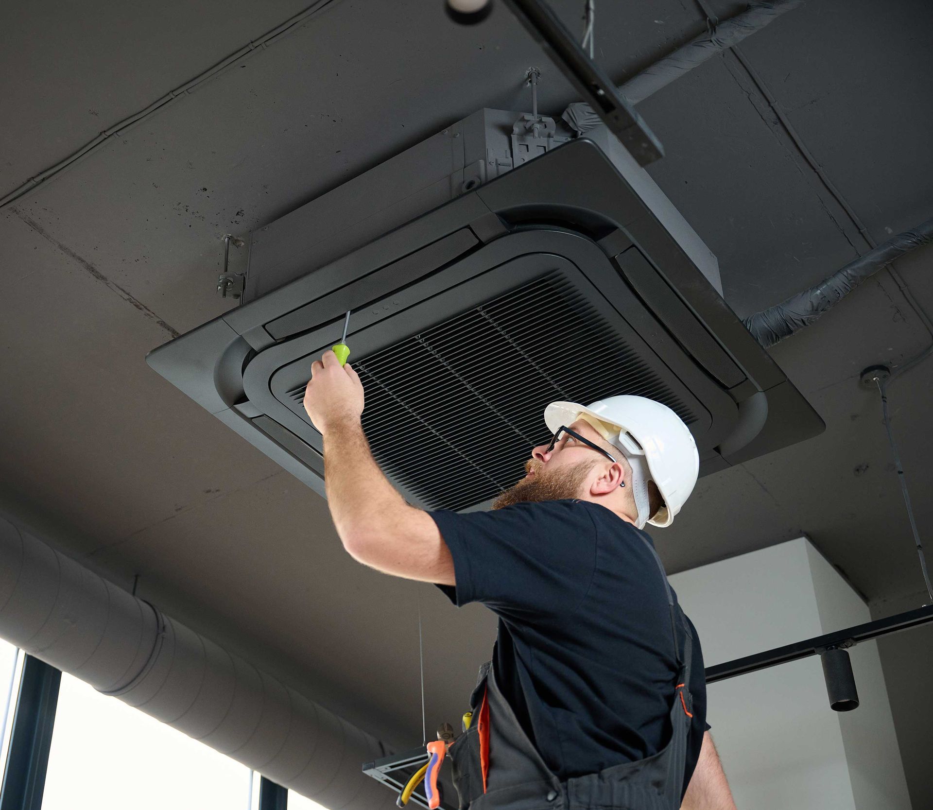 Man in a hardhat repairs ceiling air conditioning unit with a screwdriver.