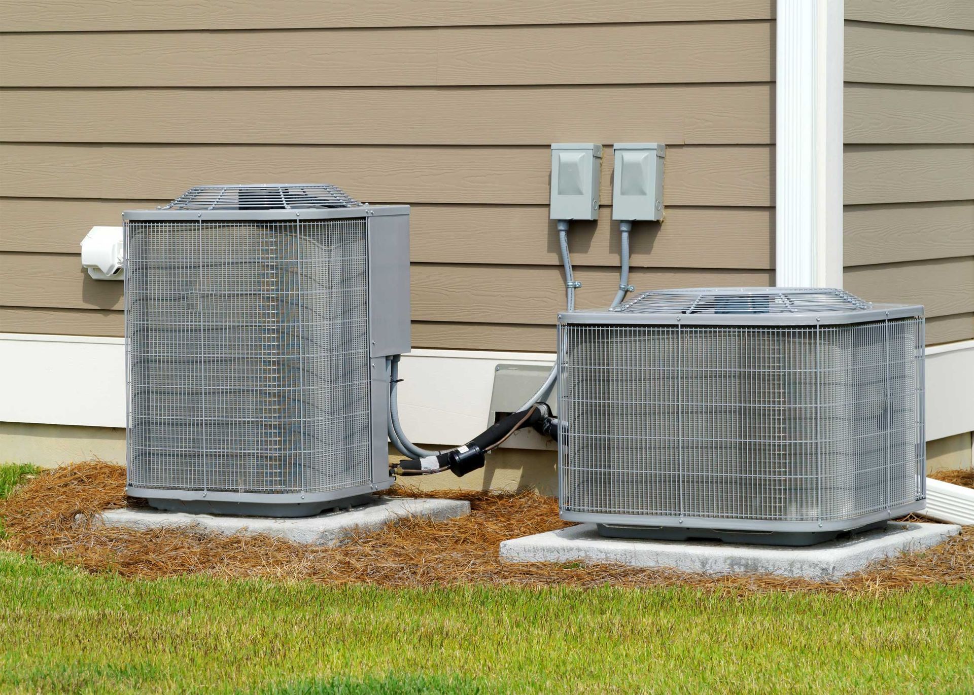 Two outdoor air conditioning units on concrete pads beside a light brown house with a green lawn.