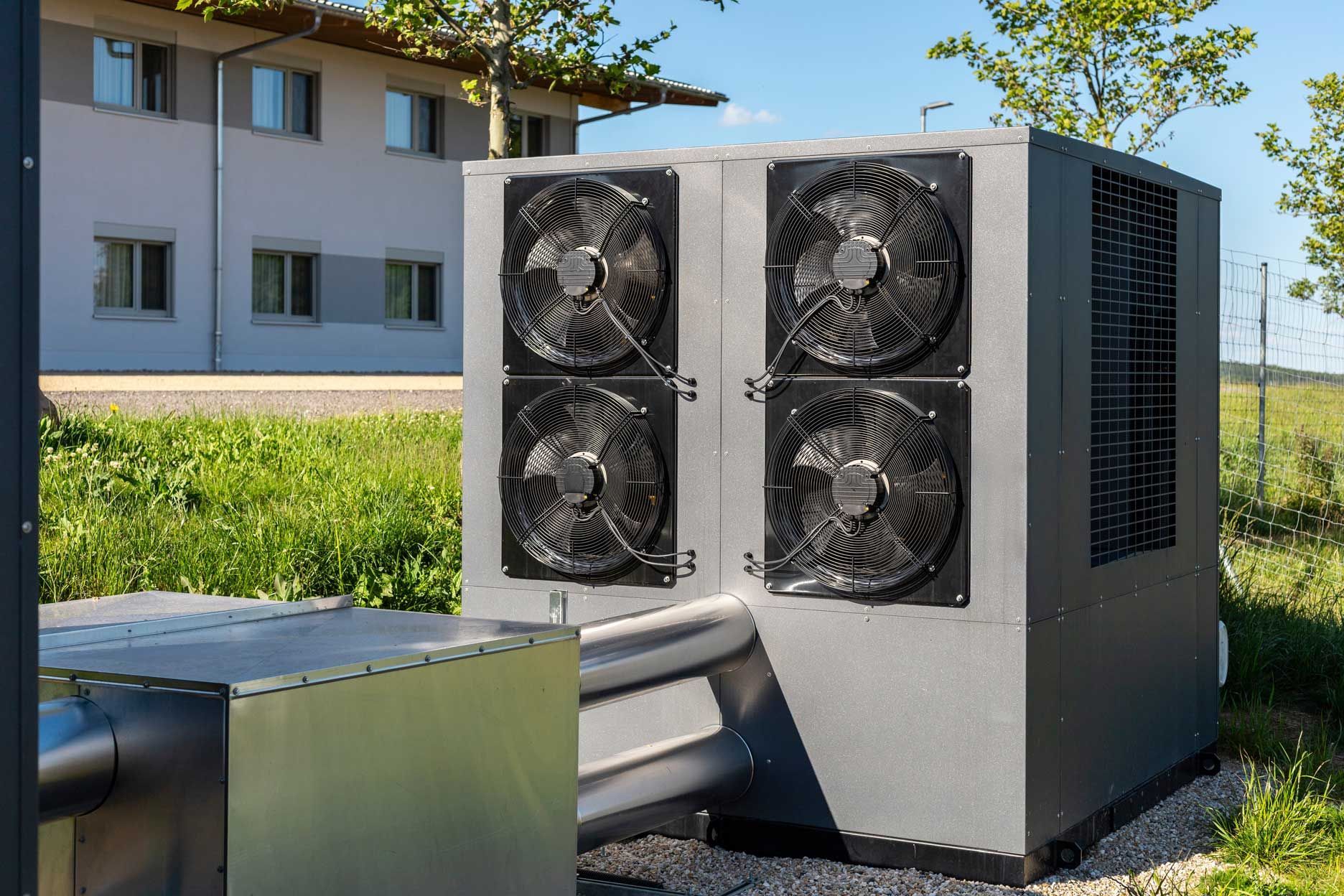 Large, gray heat pump unit with four fans outdoors near a building and grass.