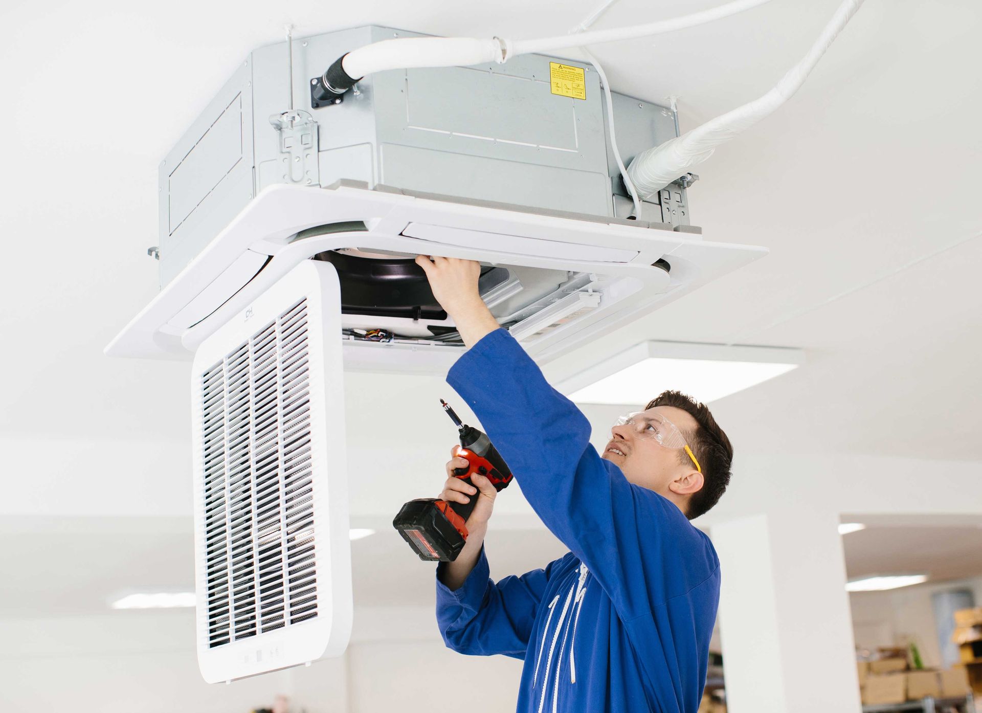 HVAC technician in blue overalls uses a drill on a ceiling-mounted air conditioning unit.