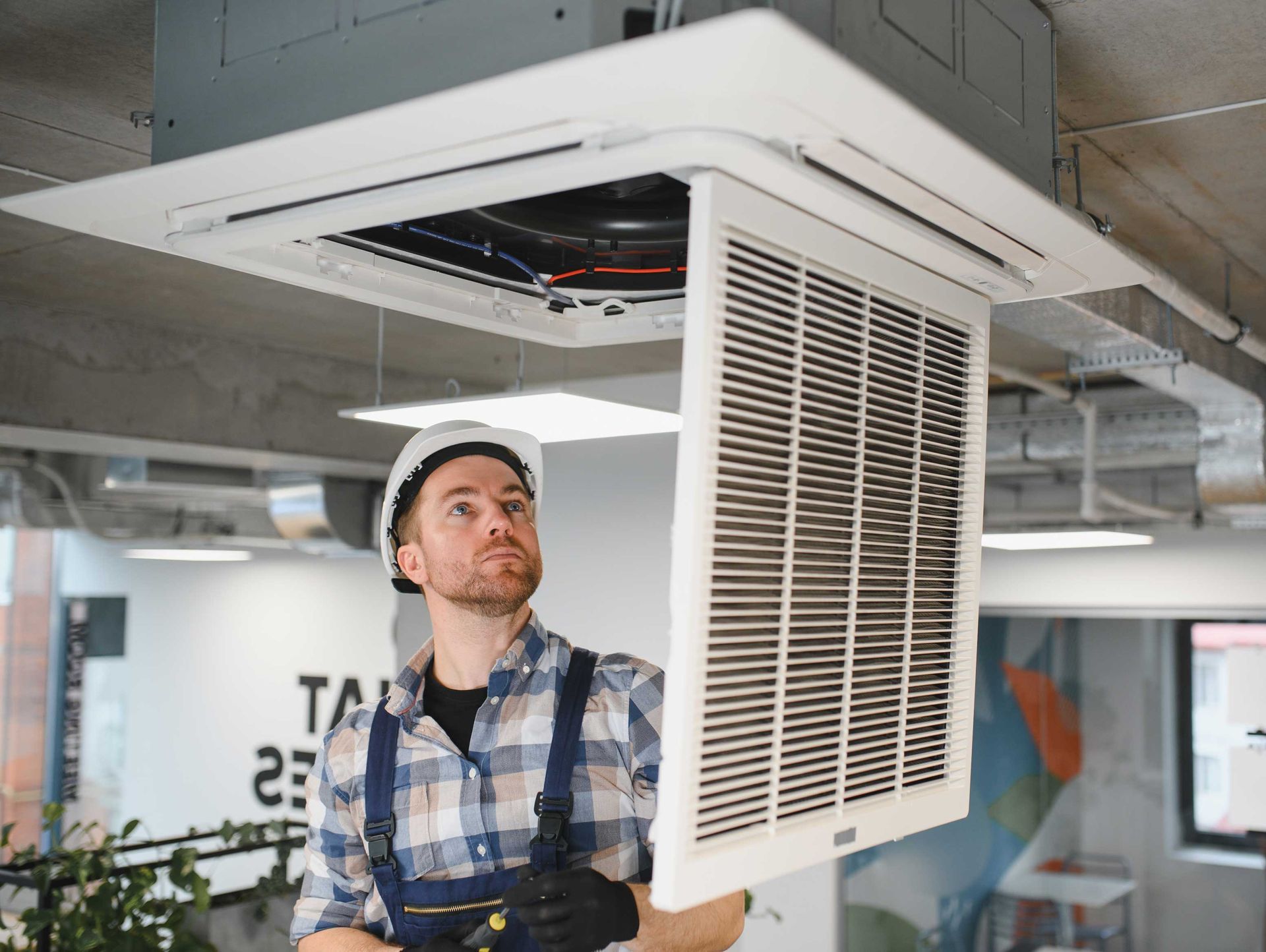 Man in hard hat inspecting ceiling air conditioning unit.