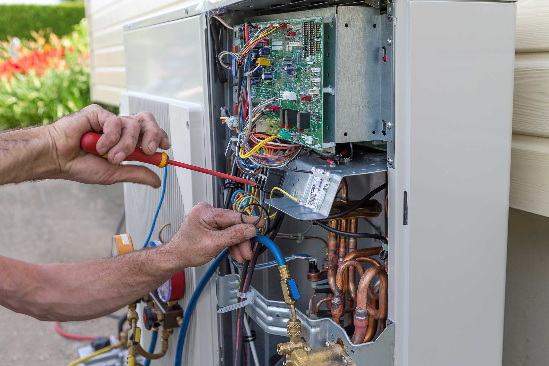 Person uses a screwdriver on the internal components of an outdoor appliance near a building and greenery.