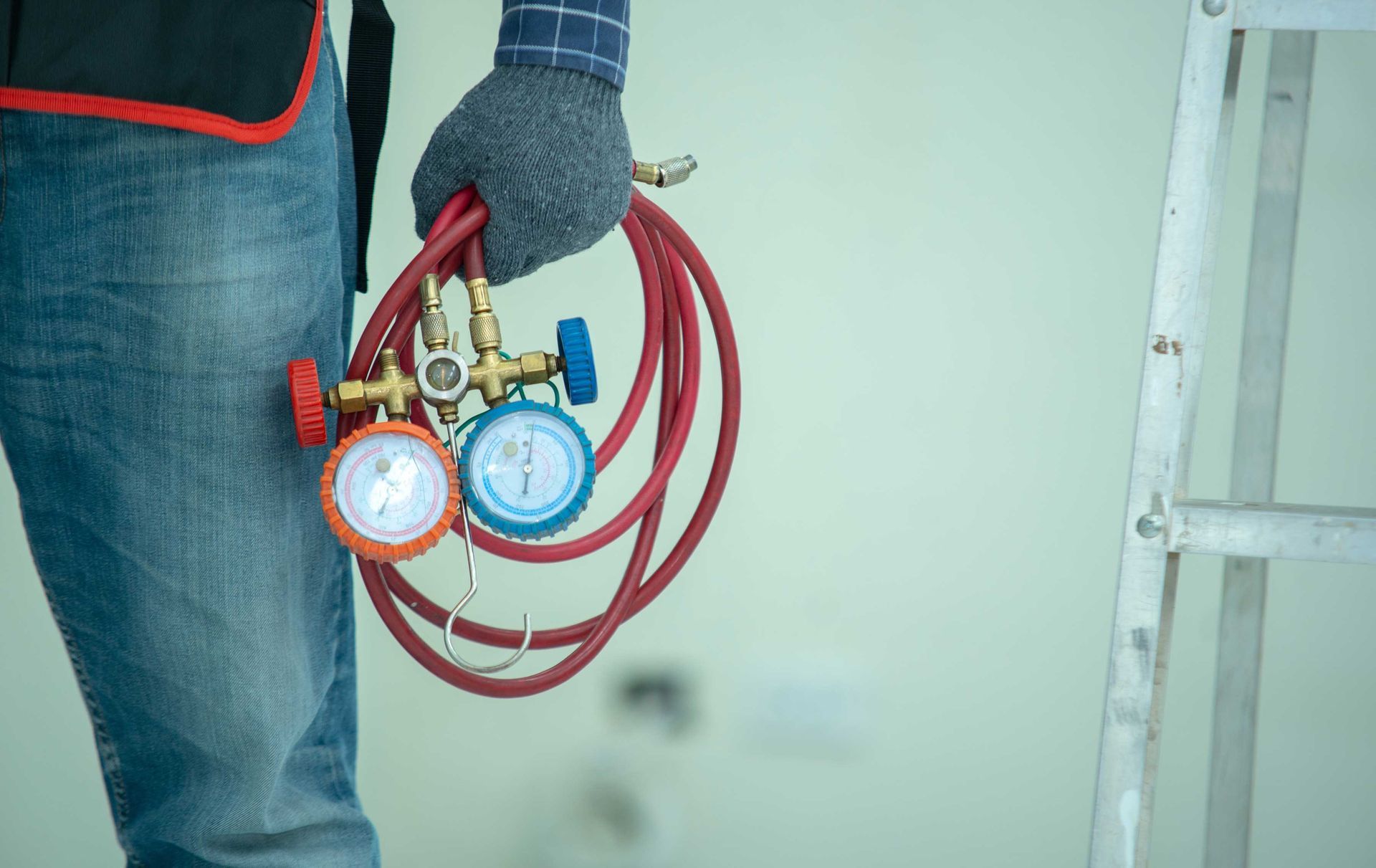 HVAC technician holding gauges and hoses near a ladder, wearing gloves and jeans.